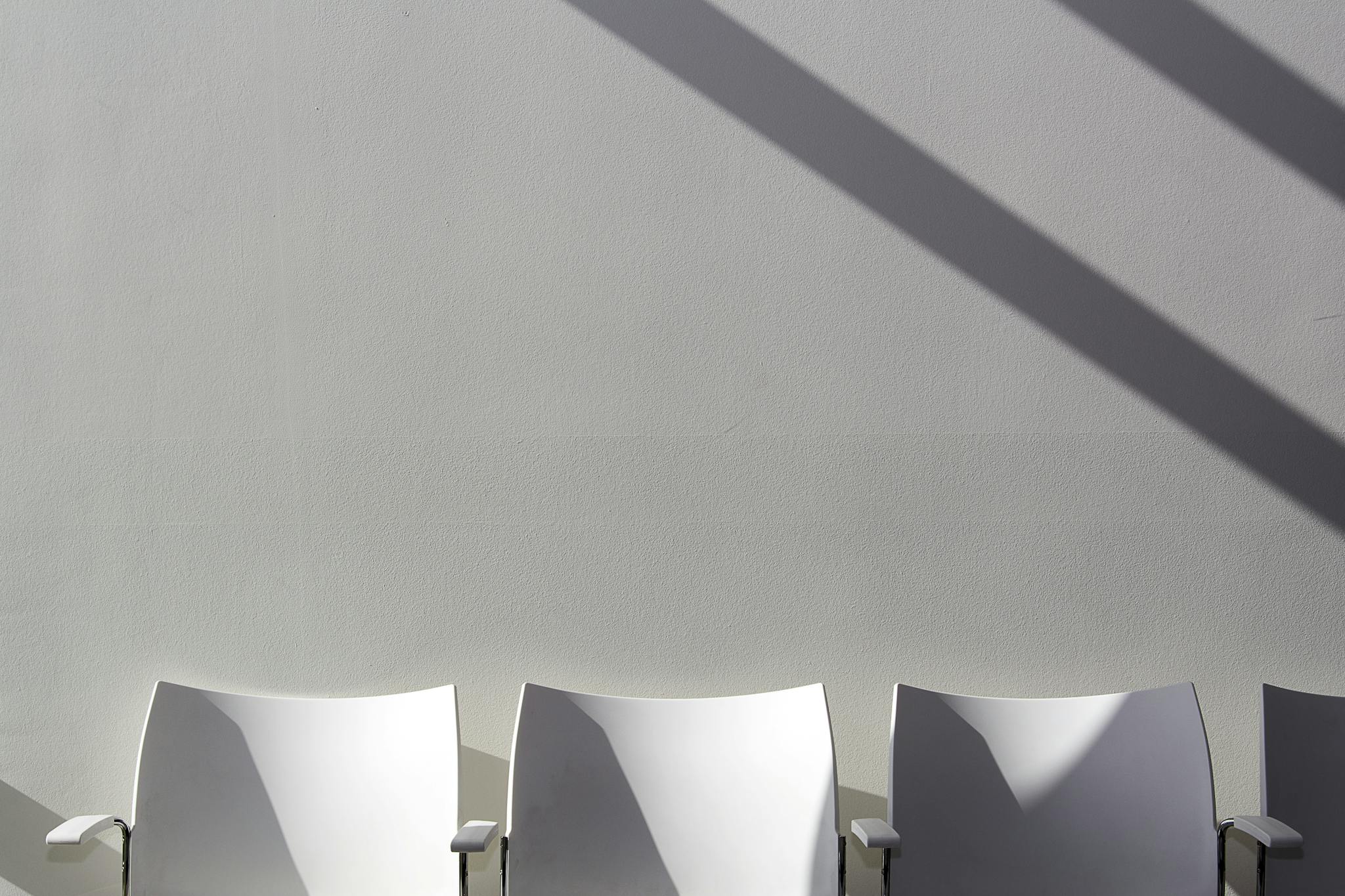 Three white chairs with long shadows against a plain wall, embodying minimalist design.